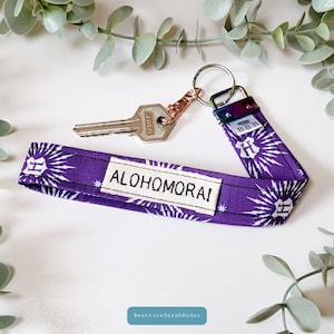 May include: Purple fabric keychain with a white design featuring the Hogwarts crest and the text "ALOHOMORA". The keychain has a silver key attached to a rose gold split ring.
