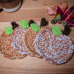 May include: Four crocheted pumpkin-shaped coasters in brown and white yarn with green yarn leaves. The coasters are arranged in a row on a wooden surface.