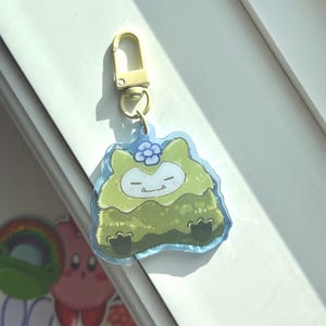 Può includere: Un portachiavi in acrilico trasparente con un personaggio dei cartoni animati in tonalità di verde, blu e bianco. Il personaggio ha gli occhi chiusi e un piccolo sorriso, con un fiore blu sulla testa. Il portachiavi ha un fermaglio color oro.