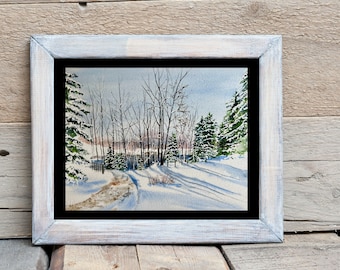May include: Watercolor painting of a snowy winter landscape, framed in a white-washed wooden frame with a black mat. The artwork depicts a path winding through snow-covered trees, with long shadows cast by the sun.