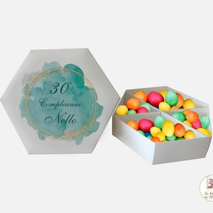 Può includere: Una scatola esagonale bianca con un design acquerellato turchese e glitter dorati. La scatola ha il testo "30° Compleanno Nello" sul coperchio. La scatola è aperta e piena di uova di cioccolato ricoperte di caramelle colorate.