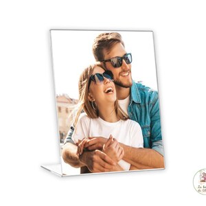 Puede incluir: Una pareja con gafas de sol sonríe y se abraza mientras mira a la cámara. El hombre lleva una camisa vaquera azul y la mujer lleva una camisa blanca.