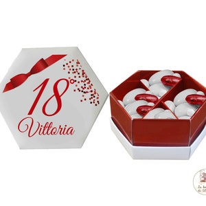 Può includere: Una scatola esagonale bianca con un nastro rosso e il testo "18 Vittoria" sulla parte superiore. La scatola è aperta e rivela una scatola esagonale rossa piena di caramelle bianche e rosse.