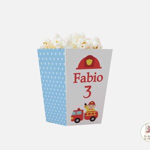 Può includere: Una scatola di popcorn blu e bianca con un design di camion dei pompieri rosso e pompiere. La scatola dice "Fabio 3" sul davanti.