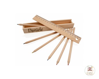 Puede incluir: Una caja de lápices de madera con el nombre "Daniele" grabado en la parte delantera. La caja contiene seis lápices de madera y una regla de madera con marcas hasta 20 cm.