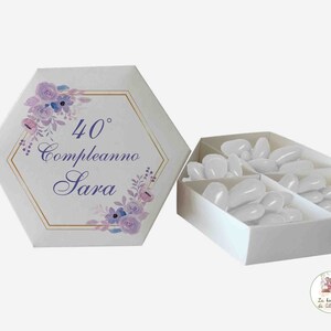 Può includere: Scatola esagonale bianca con un design floreale e dettagli dorati. La scatola ha il testo "40° compleanno Sara" sul davanti. La scatola è aperta e piena di caramelle bianche.