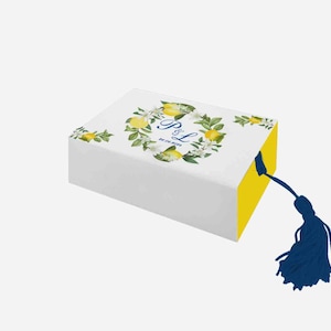 Op de afbeelding: Een witte cadeaudoos met een geel deksel en een blauwe kwast. De doos is versierd met een bloemmotief met citroenen en witte bloemen. De tekst "P&L" is op de doos gedrukt.