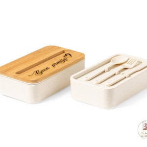 Op de afbeelding: Een witte en beige lunchbox met een bamboe deksel. Het deksel heeft de tekst "Buon Pranzo" gegraveerd. De lunchbox wordt geleverd met een bijpassend bestekset, inclusief een vork, mes en lepel.