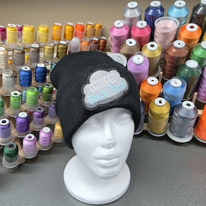 May include: A black knit beanie with a white and light blue embroidered patch that reads "CERTIFIED Black Cloud." The beanie is displayed on a white foam head, with spools of colorful thread in the background.