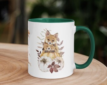 May include: White ceramic mug with a green rim. The mug features a watercolour illustration of two foxes nestled together inside a pumpkin. The foxes are surrounded by leaves and flowers.