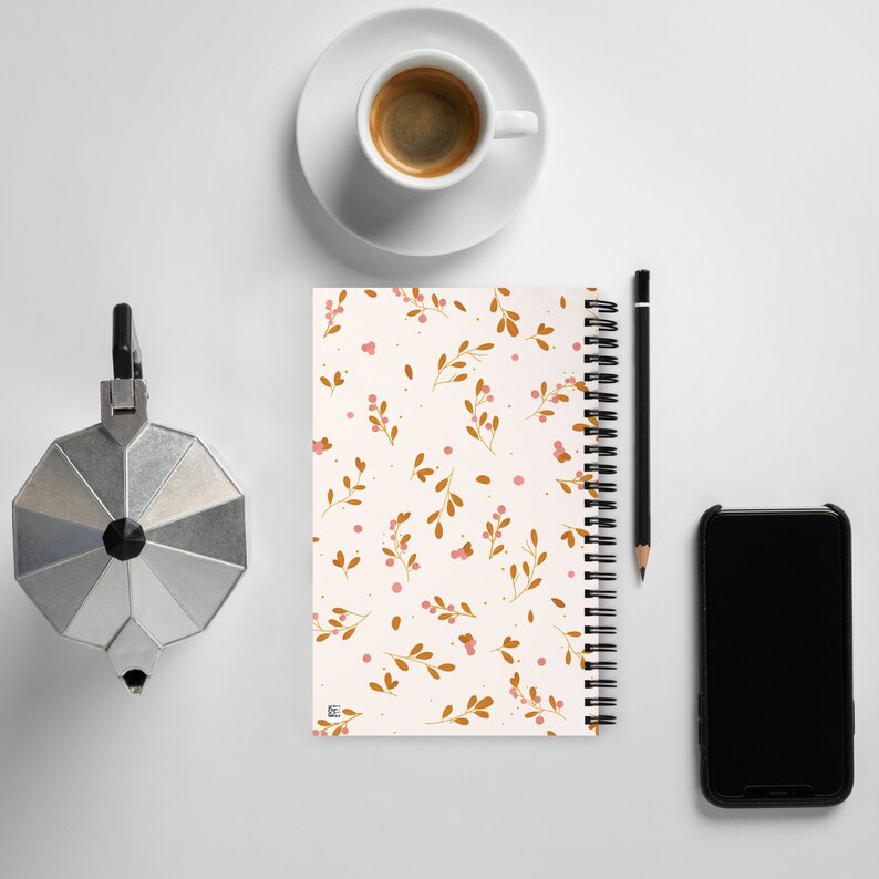 May include: A white spiral bound notebook with a floral pattern. The pattern features small pink berries and brown branches on a white background. The notebook is lying flat on a white surface with a cup of coffee, a pencil, and a black smartphone beside it.