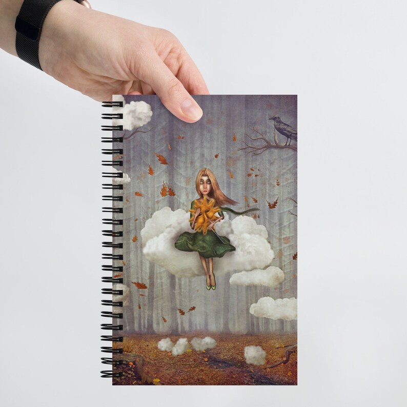 May include: A spiral bound notebook featuring a colourful illustration of a girl sitting on a cloud in a forest. The girl is holding a yellow flower and wearing a green dress. The background is a misty forest with falling leaves and a crow perched on a branch.