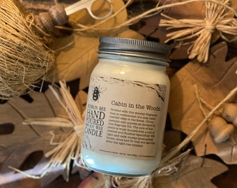 May include: A glass jar candle with a silver lid and a brown label that reads "Cabin in the Woods". The label has a small illustration of a bee and text that says "Bumblin Bee Hand Poured Soy Wax Candle".