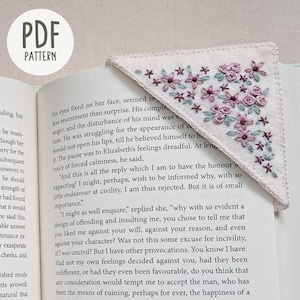 May include: A white felt bookmark with a floral embroidery design in shades of pink and green. The bookmark is folded into a triangle shape and is placed on top of an open book.