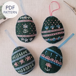 Könnte beinhalten: Vier Filz-Ostereier mit verschiedenen Stickmustern. Jedes Ei hat eine Schleifenöse zum Aufhängen. Die Eier sind grün mit rosa, blauen und weißen Stickereien. Die Muster umfassen Blumen, Blätter und geometrische Muster.
