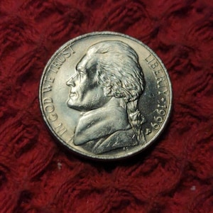 Op de afbeelding: Een zilveren Amerikaanse nikkelmunt met het profiel van Thomas Jefferson op de voorzijde. De munt heeft de tekst "IN GOD WE TRUST" en "LIBERTY 1965".