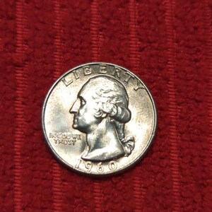 Könnte beinhalten: Eine silberne 1960er Vierteldollarmünze mit dem Profil von George Washington nach links. Die Münze trägt die Inschriften "LIBERTY" und "IN GOD WE TRUST". Auf rotem Hintergrund.