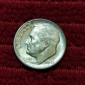 May include: A silver US dime coin with the year 1946 inscribed on the side. The coin features a profile of a man with the word "LIBERTY" inscribed above his head and "IN GOD WE TRUST" inscribed below.