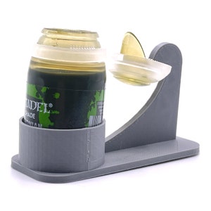 May include: A gray 3D printed paint bottle holder with a clear bottle of green paint inside. The bottle has a label that says "Citadel Shade" and "Nurgling Green".