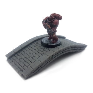 May include: A grey stone bridge miniature for tabletop gaming. The bridge is arched and has a textured surface. A red painted miniature figure stands on the bridge.
