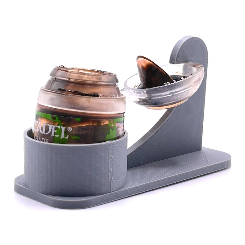 May include: A gray 3D printed paint pot holder with a Citadel paint pot in a holder and a separate lid holder. The paint pot is brown and has the word "Citadel" on it.