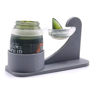 May include: A gray 3D printed paint pot holder with a clear plastic paint mixing tray. The holder has a slot for a paint bottle and a small lip to hold the mixing tray. The paint bottle is green and has the label "Citadel Colour".
