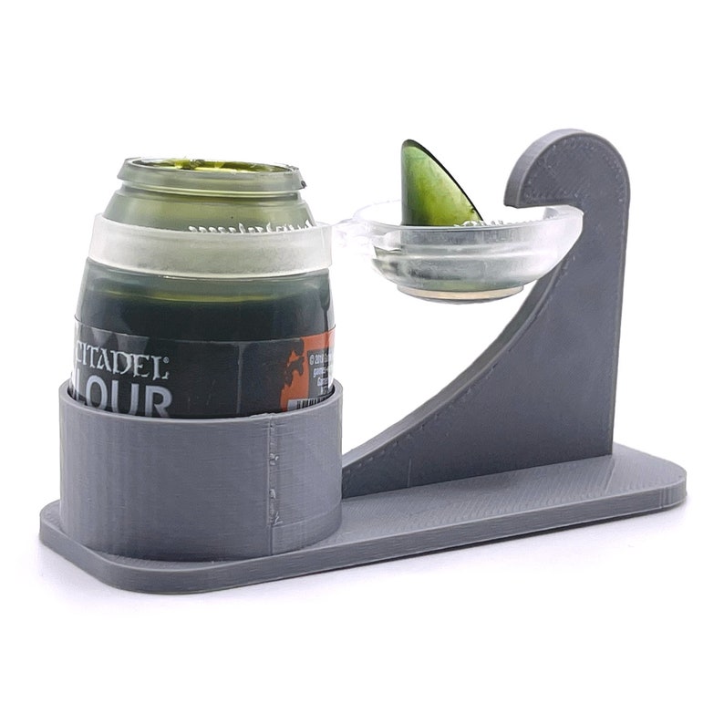 May include: A gray 3D printed paint pot holder with a clear plastic paint mixing tray. The holder has a slot for a paint bottle and a small lip to hold the mixing tray. The paint bottle is green and has the label "Citadel Colour".