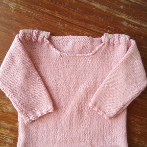 Può includere: Un maglione rosa chiaro lavorato a mano con maniche lunghe. Il maglione presenta uno scollo tondo e piccoli bottoni decorativi sulle spalle. Il fondo e i polsini delle maniche hanno un bordo smerlato. Il maglione è realizzato con un filato morbido e strutturato.