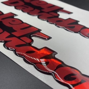Op de afbeelding: Twee rode en zwarte 3D vinyl stickers met het woord "Revolution" in een vet, gestileerd lettertype.