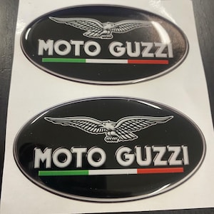 Może przedstawiać: Dwa czarne owalne naklejki z białym orłem i tekstem "MOTO GUZZI" białymi literami. Naklejki mają również zielono-biało-czerwony pasek, przypominający flagę Włoch.