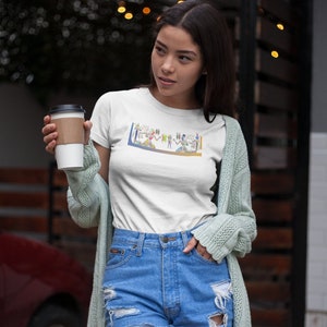 May include: A white t-shirt with a colorful graphic print of ancient Egyptian hieroglyphs. The shirt is being worn by a person with long brown hair and is paired with blue denim shorts and a light green cardigan.
