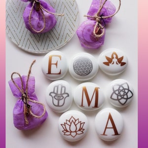 May include: Eight white ceramic stones with gold accents, each featuring a different design. The designs include the letters E, M, A, a lotus flower, a hamsa hand, a geometric pattern, a yoga pose, and a flower of life. The stones are arranged in a circle on a white surface.