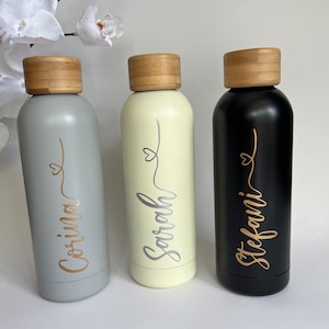 May include: Three stainless steel water bottles with bamboo lids. The bottles are grey, cream, and black. Each bottle has a name written on it in gold or silver lettering: Corina, Sarah, and Stefani.