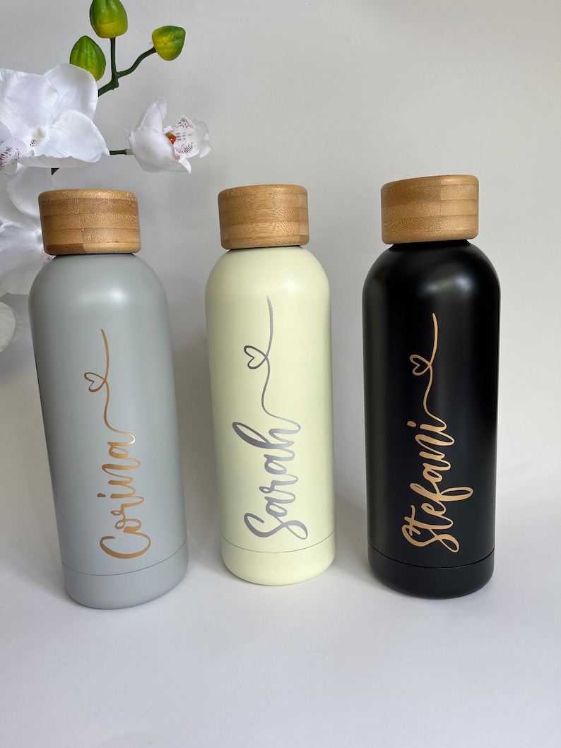 May include: Three stainless steel water bottles with bamboo lids. The bottles are grey, cream, and black. Each bottle has a name written on it in gold or silver lettering: Corina, Sarah, and Stefani.