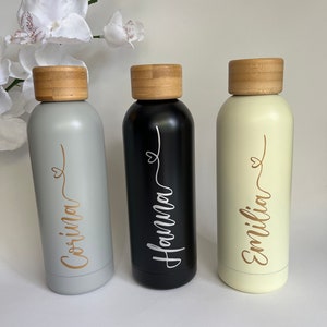 May include: Three personalized stainless steel water bottles with bamboo lids. The bottles are gray, black, and cream, and each has a gold name written on it: Corina, Hanna, and Emilia.