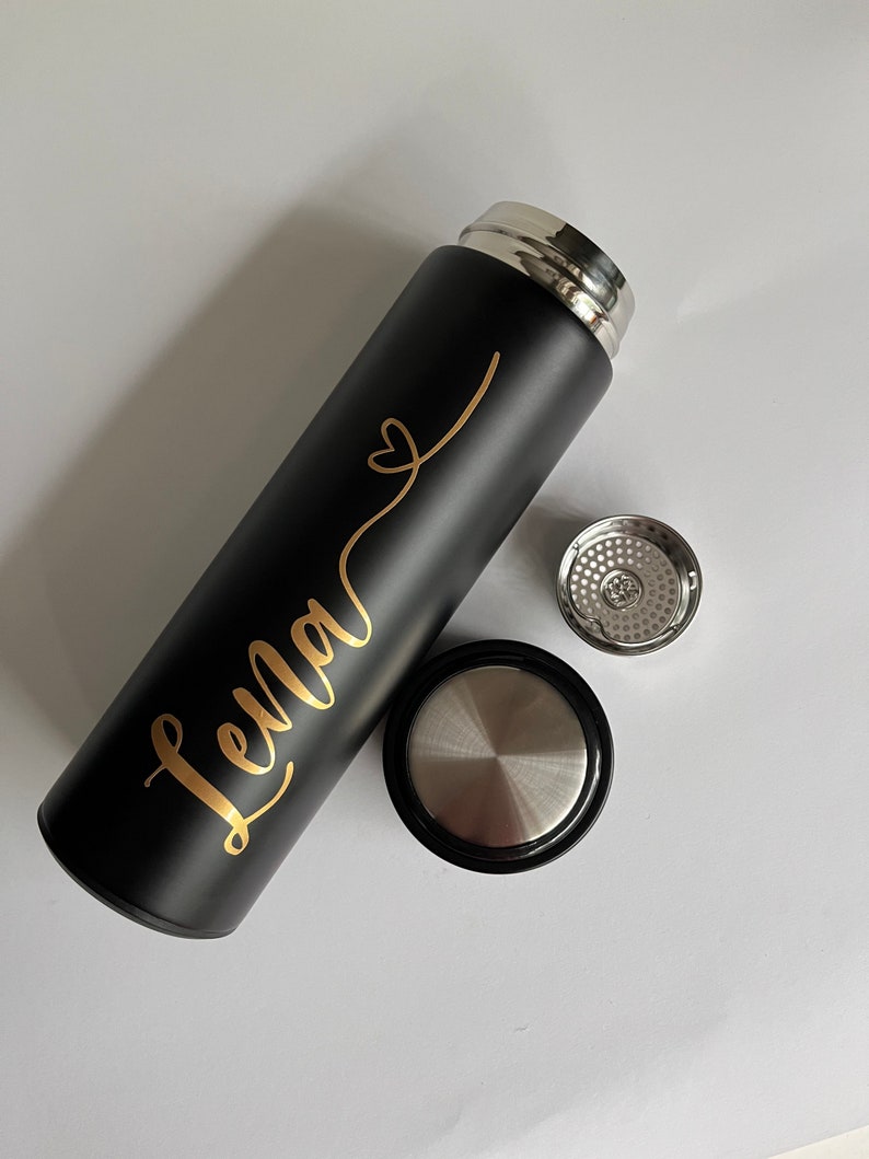May include: Black stainless steel water bottle with a gold inscription that reads "Lena" with a heart symbol. The bottle has a black lid and a silver lid with a strainer.