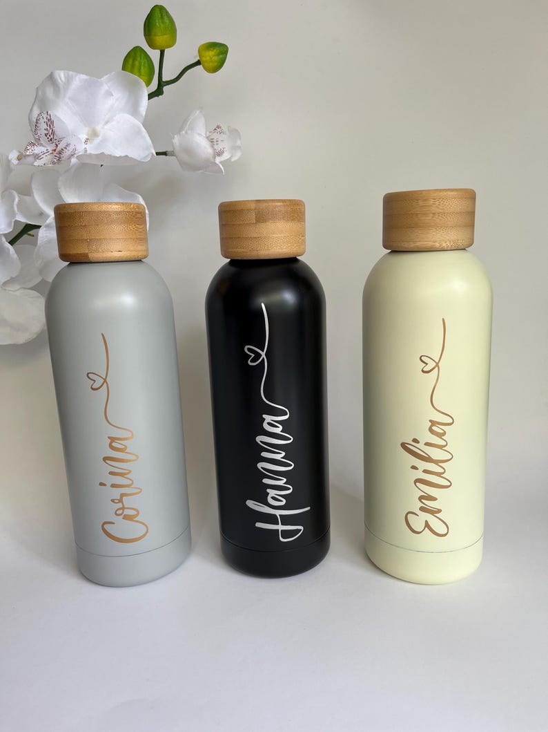 May include: Three personalized water bottles with bamboo lids. The bottles are gray, black, and cream with gold lettering. The names on the bottles are Corina, Hanna, and Emilia.