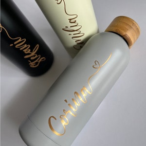 May include: Two stainless steel water bottles with bamboo lids. One bottle is gray with gold lettering that says "Corinna" and the other is white with gold lettering that says "Emilia".
