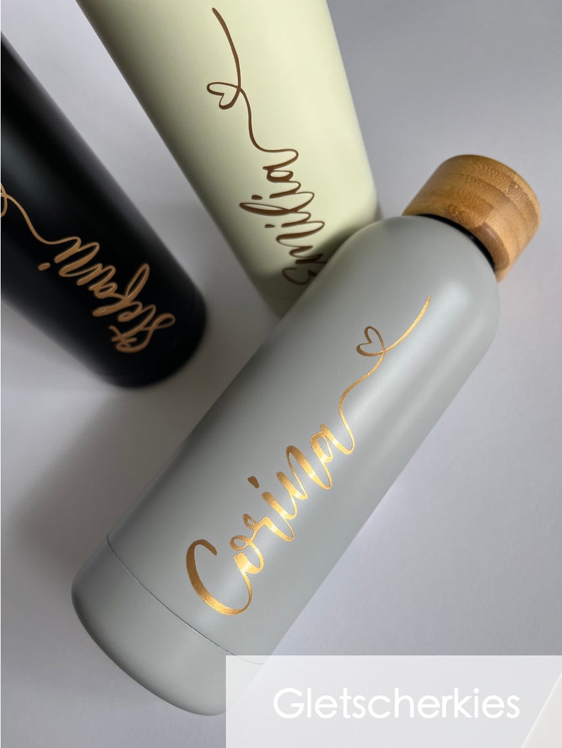 May include: Two stainless steel water bottles with bamboo lids. One bottle is gray with gold lettering that says "Corinna" and the other is white with gold lettering that says "Emilia".