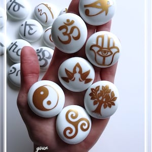May include: A hand holding a collection of white stones with gold symbols. The symbols include the Om symbol, the Eye of Horus, the Hamsa hand, a tree of life, a yin yang symbol, a lotus flower, and a triskelion. The stones are smooth and polished.