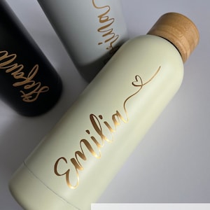 May include: A light green stainless steel water bottle with a wooden lid and a gold inscription that reads "Emilia" with a small heart. The bottle is on a white surface.