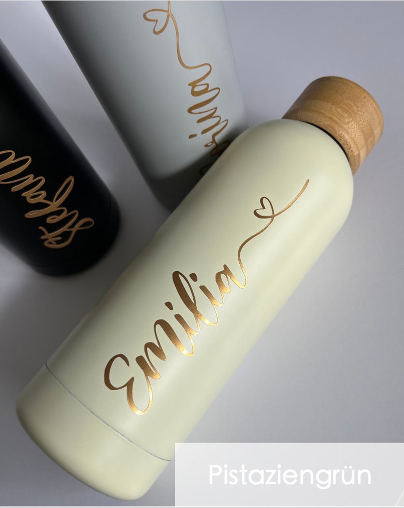 May include: A light green stainless steel water bottle with a wooden lid and a gold inscription that reads "Emilia" with a small heart. The bottle is on a white surface.