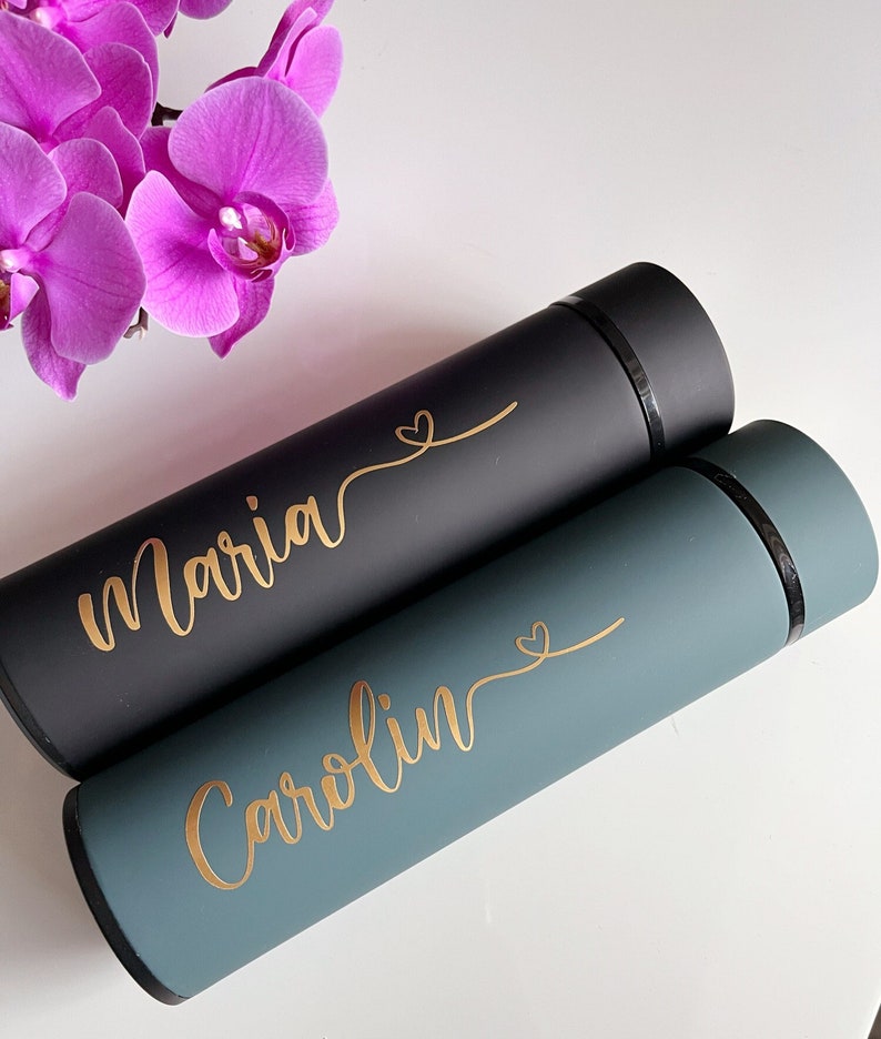 May include: Two personalized water bottles, one black with the name "Maria" and one teal with the name "Carolyn". Both bottles have a gold heart design.