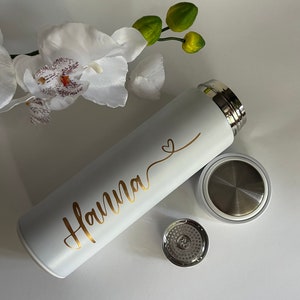 May include: White stainless steel water bottle with a gold inscription that reads "Hanna" and a small heart symbol. The bottle has a matching lid with a built-in strainer.