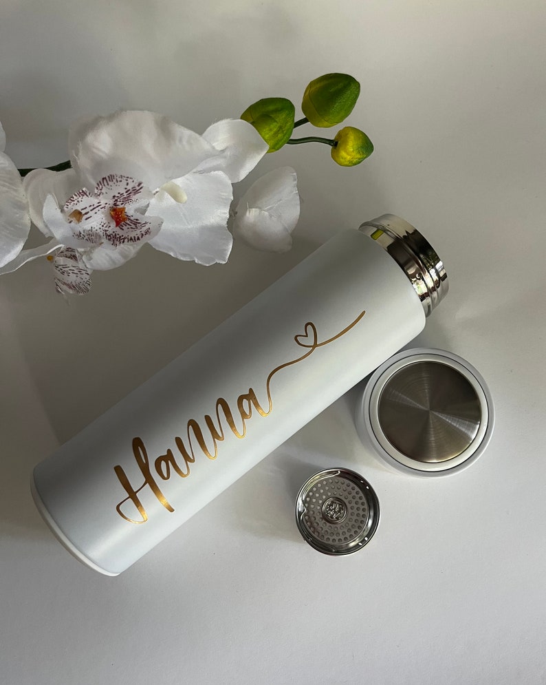 May include: White stainless steel water bottle with a gold inscription that reads "Hanna" and a small heart symbol. The bottle has a matching lid with a built-in strainer.