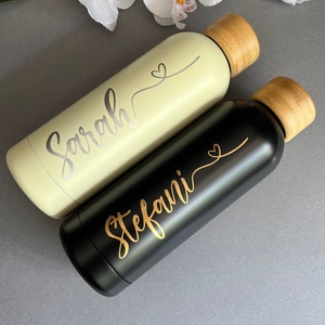 May include: Two personalized water bottles with bamboo lids. One bottle is light green with the name "Sarah" and a heart design. The other bottle is black with the name "Stefani" and a heart design.