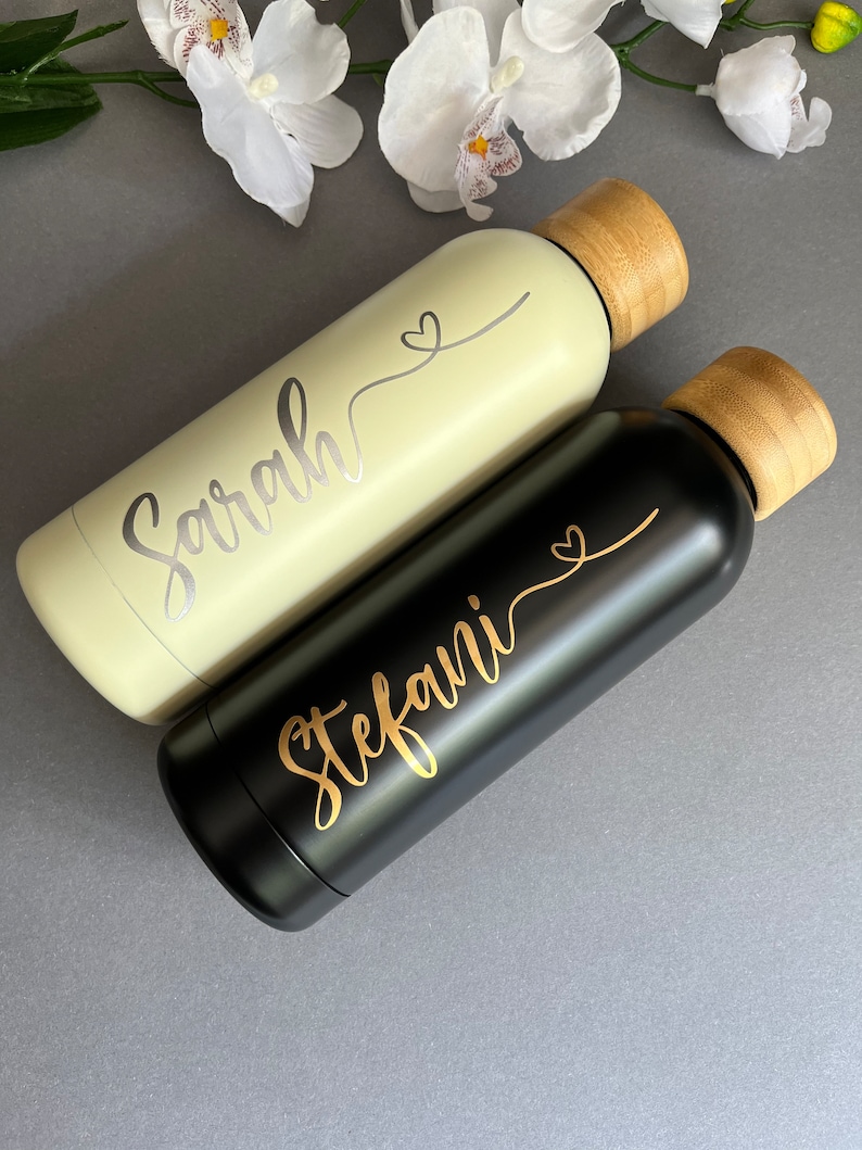 May include: Two personalized water bottles with bamboo lids. One bottle is light green with the name "Sarah" and a heart design. The other bottle is black with the name "Stefani" and a heart design.