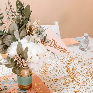 Puede incluir: Un arreglo floral blanco en un jarr&oacute;n dorado y turquesa con un mantel de confeti rosa y dorado y un letrero rosa que dice "Disney World".