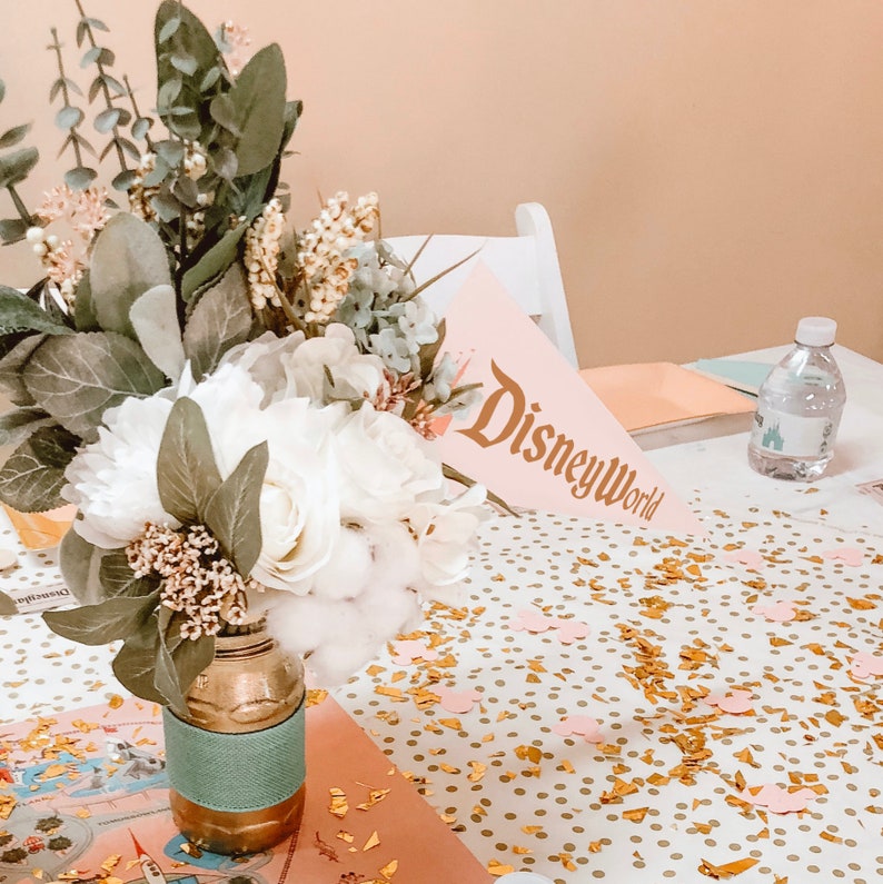 Puede incluir: Un arreglo floral blanco en un jarr&oacute;n dorado y turquesa con un mantel de confeti rosa y dorado y un letrero rosa que dice "Disney World".