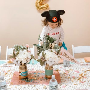 Puede incluir: Una mesa preparada para una fiesta de cumplea&ntilde;os con tem&aacute;tica de Disney World. La mesa est&aacute; cubierta con un mantel blanco con confeti dorado. Hay dos centros de mesa florales en tarros de vidrio con detalles dorados y turquesa. Los tarros est&aacute;n etiquetados con "Disney". Un bander&iacute;n rosa y azul con el texto "Disney World" est&aacute; sobre la mesa.